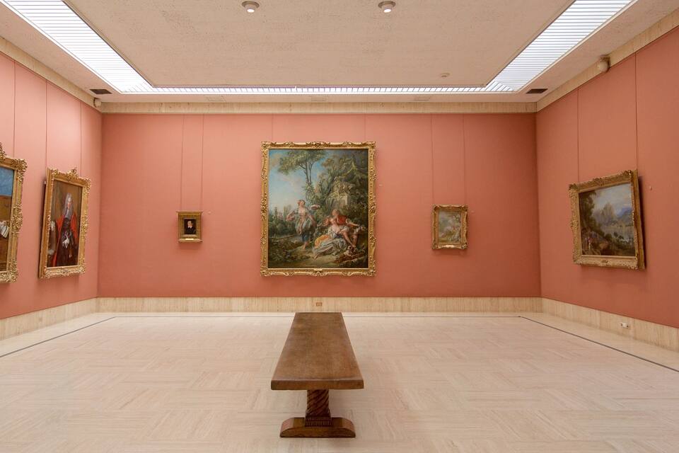 Una galería de arte con paredes y techo rosados, que exhibe pinturas clásicas y una mesa central.