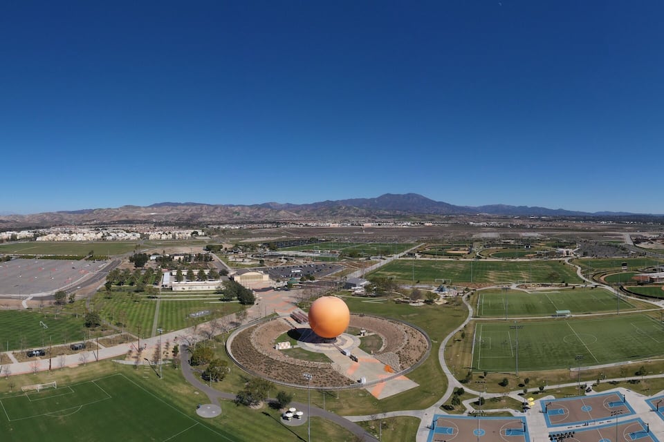 An aerial view of a sports complex with multiple fields, a large stadium, and a mountain in the background.