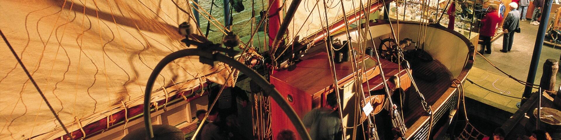 Cubierta de barco con gente de pie y sentada, cuerdas y una zona con redes.