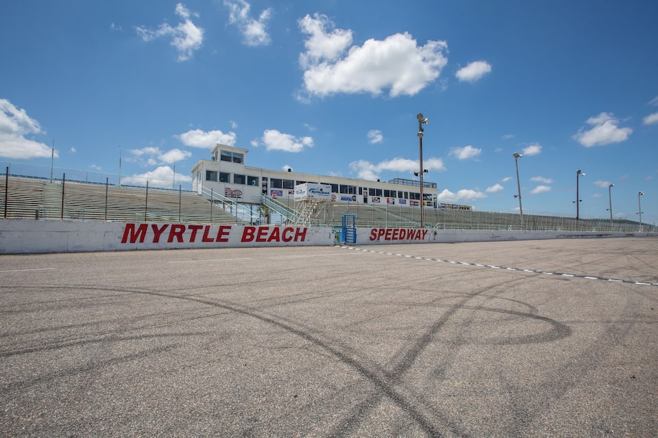 Una pista de carreras con señalización que dice 'MYRTLE BEACH SPEEDWAY', un graderío y un cielo azul despejado.
