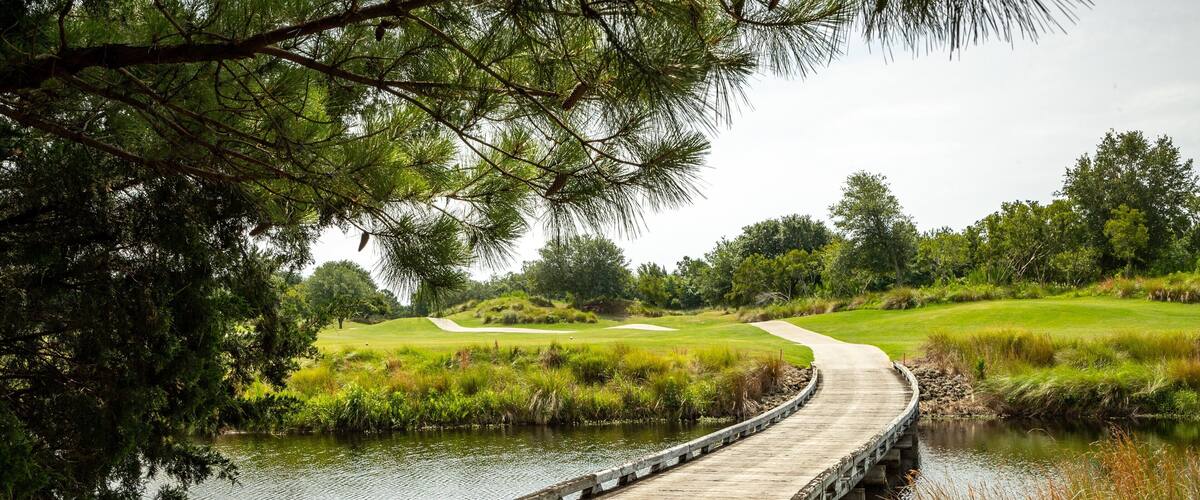 A wooden bridge over a calm river, surrounded by lush greenery and a golf course in the background.