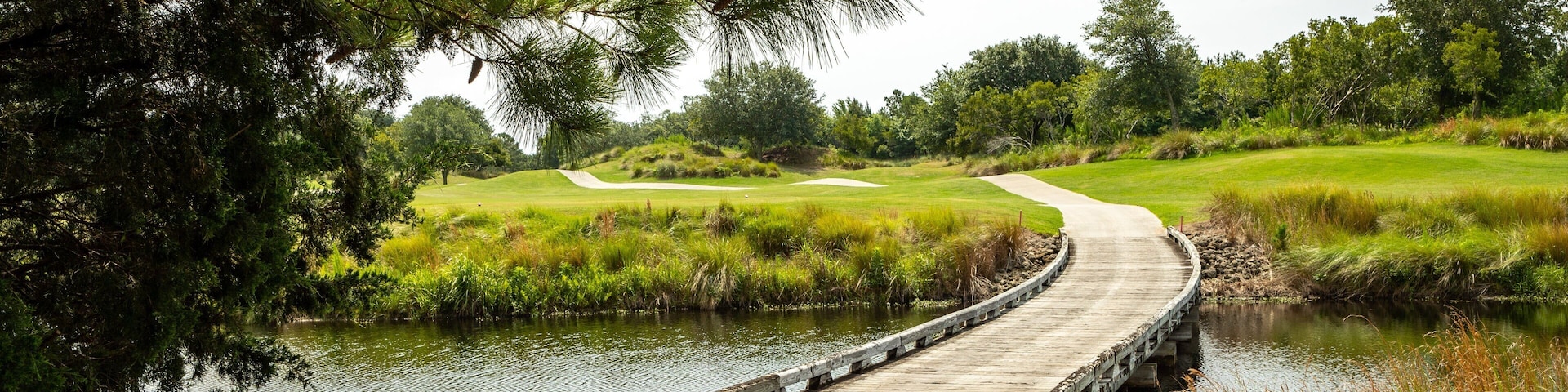 A wooden bridge over a calm river, surrounded by lush greenery and a golf course in the background.