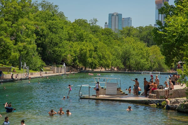 Une piscine publique entourée d’arbres verdoyants et, en arrière-plan, le paysage urbain de la ville.