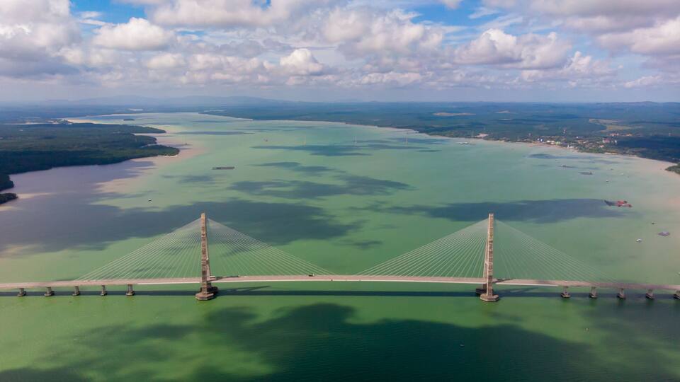 An aerial view of a bridge spanning a large body of water with a distant shoreline and cloudy sky.