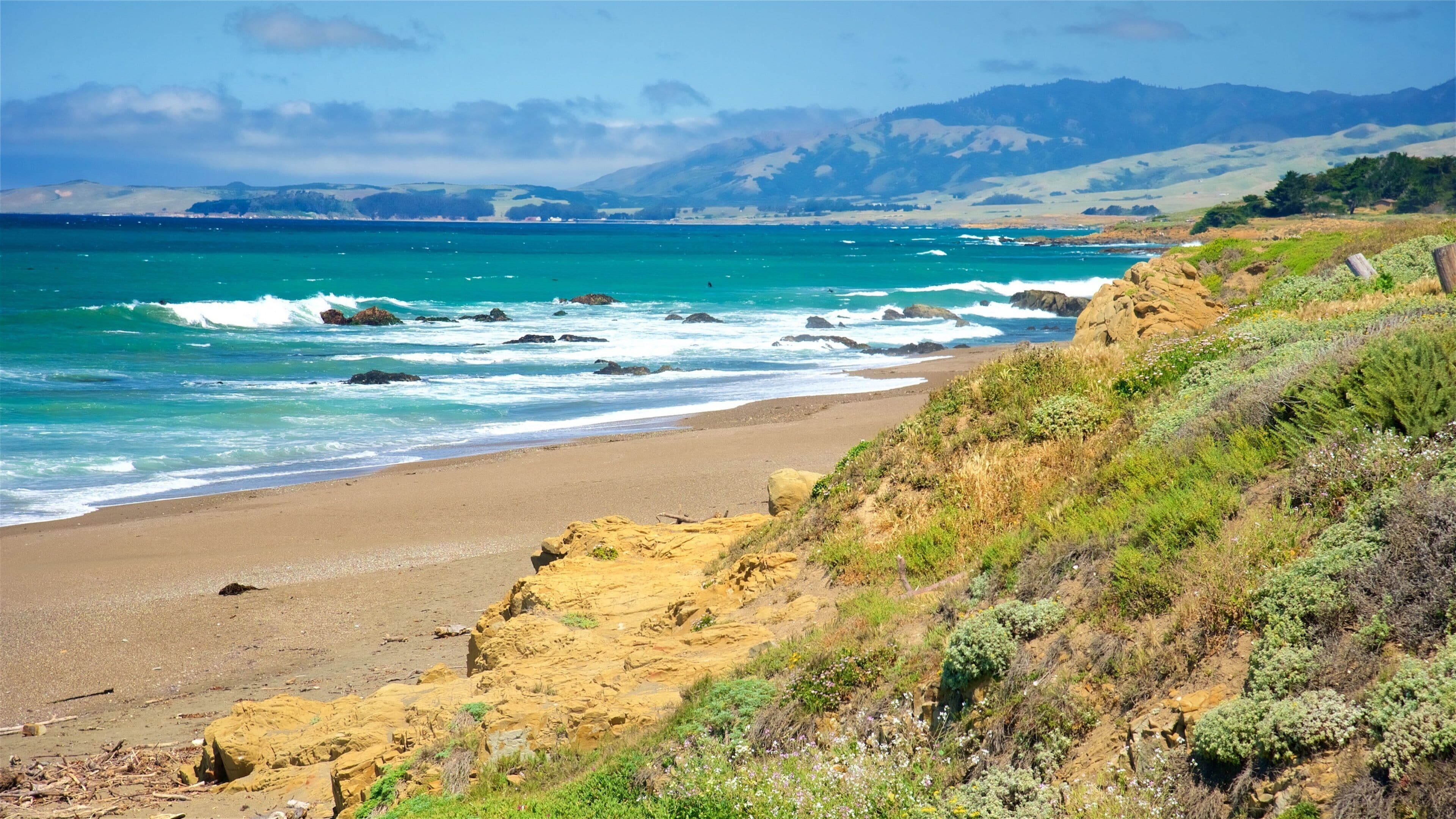 Paisaje costero con playa arenosa, acantilados rocosos y vista al océano con olas rompiendo en la orilla.