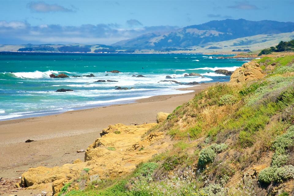 A coastal landscape with a sandy beach, rocky cliffs, and a view of the ocean with waves crashing onto the shore.