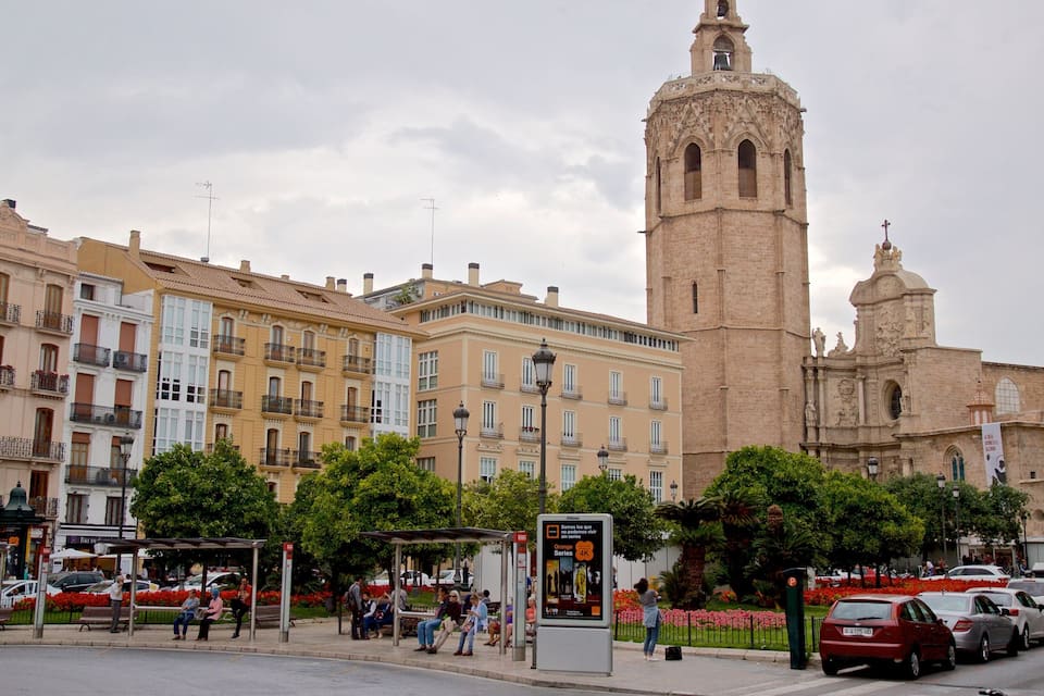 A historic tower with intricate architecture, a row of colorful buildings, and a busy street with cars and pedestrians.