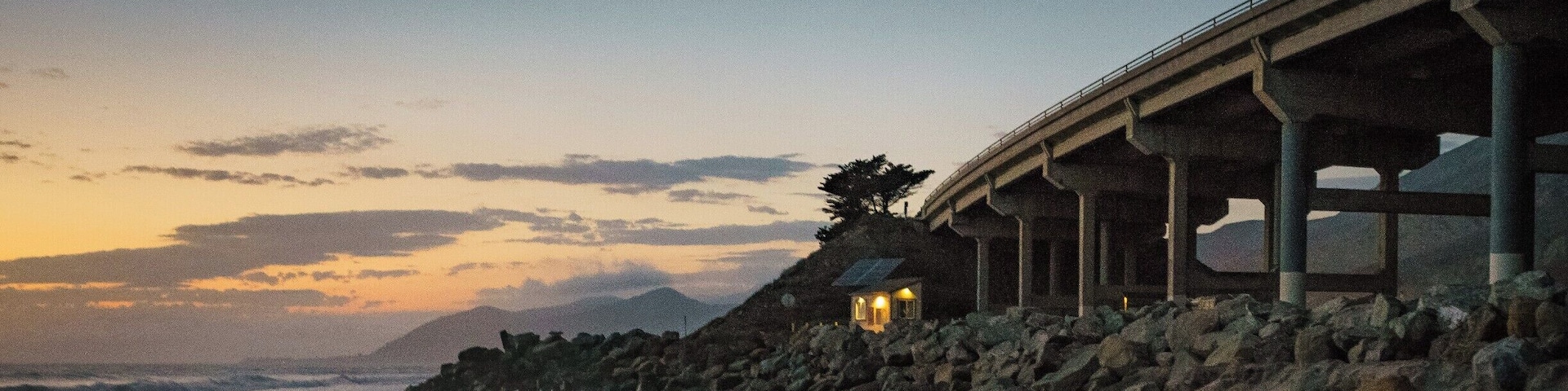 A coastal sunset with a beach, ocean waves, a bridge, and a small illuminated structure.