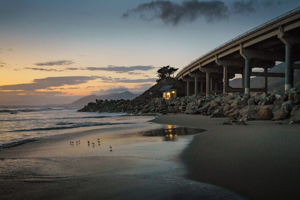 A coastal sunset with a beach, ocean waves, a bridge, and a small illuminated structure.