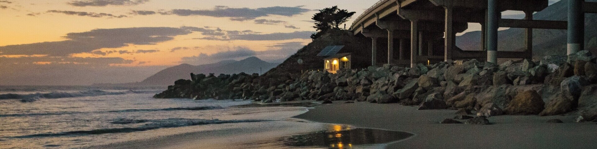 Um pôr do sol costeiro com praia, ondas do oceano, uma ponte e uma pequena estrutura iluminada.