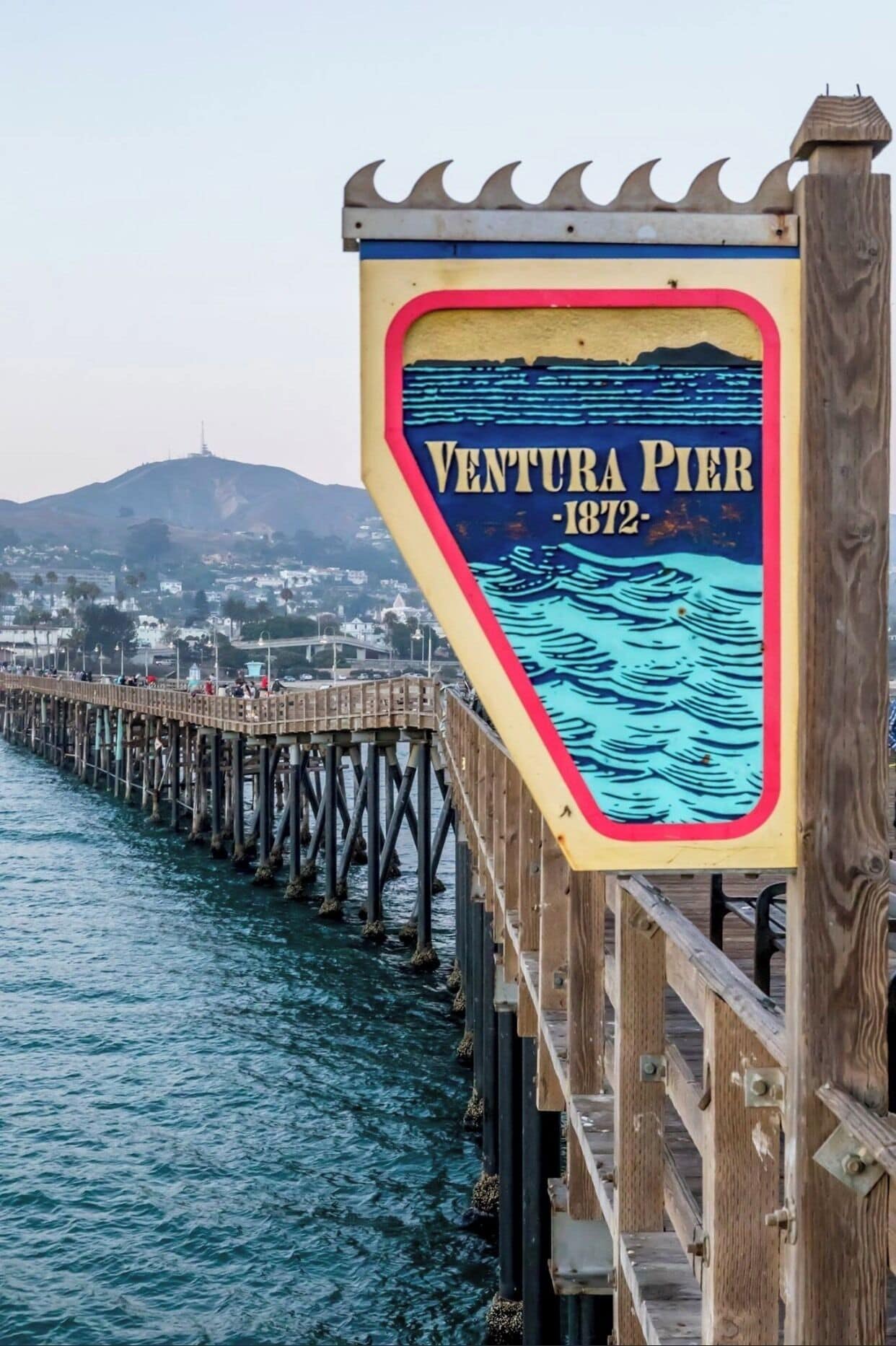 Un muelle de madera con un letrero que dice "Ventura Pier 1872" frente a un pueblo costero y montañas.
