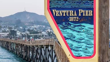 Um píer de madeira com uma placa que diz "Ventura Pier 1872", com uma paisagem de uma cidade costeira e montanhas ao fundo.