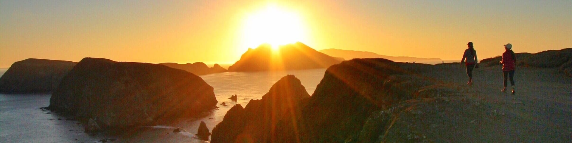 A sunset over a rocky coastline with two people walking on the beach.