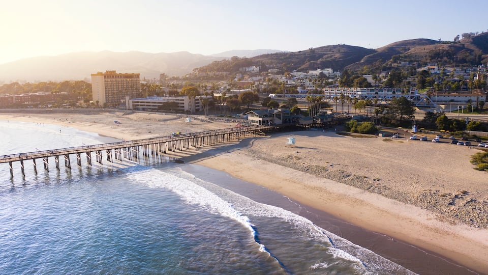 A beach with a pier extending into the ocean, a sandy shore, and buildings in the background.