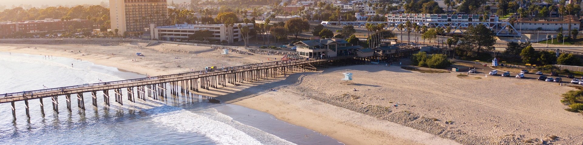 A beach with a pier extending into the ocean, a sandy shore, and buildings in the background.