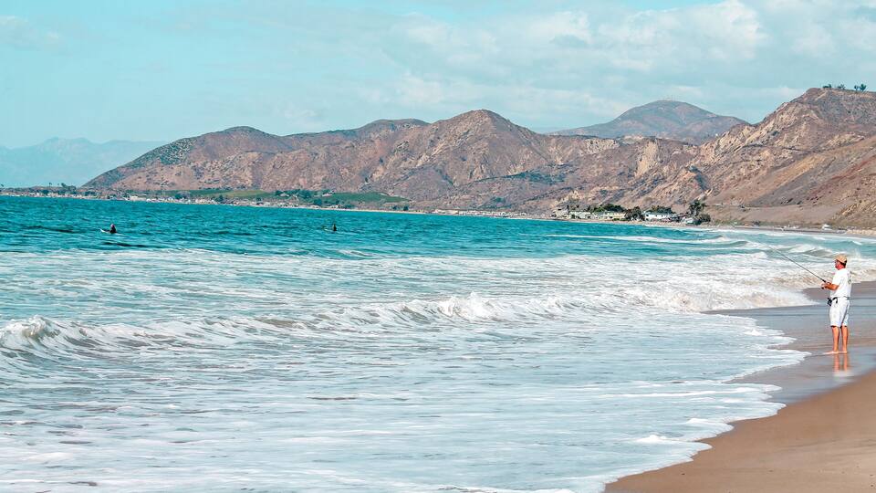A beach with waves, a person fishing, and mountains in the background.