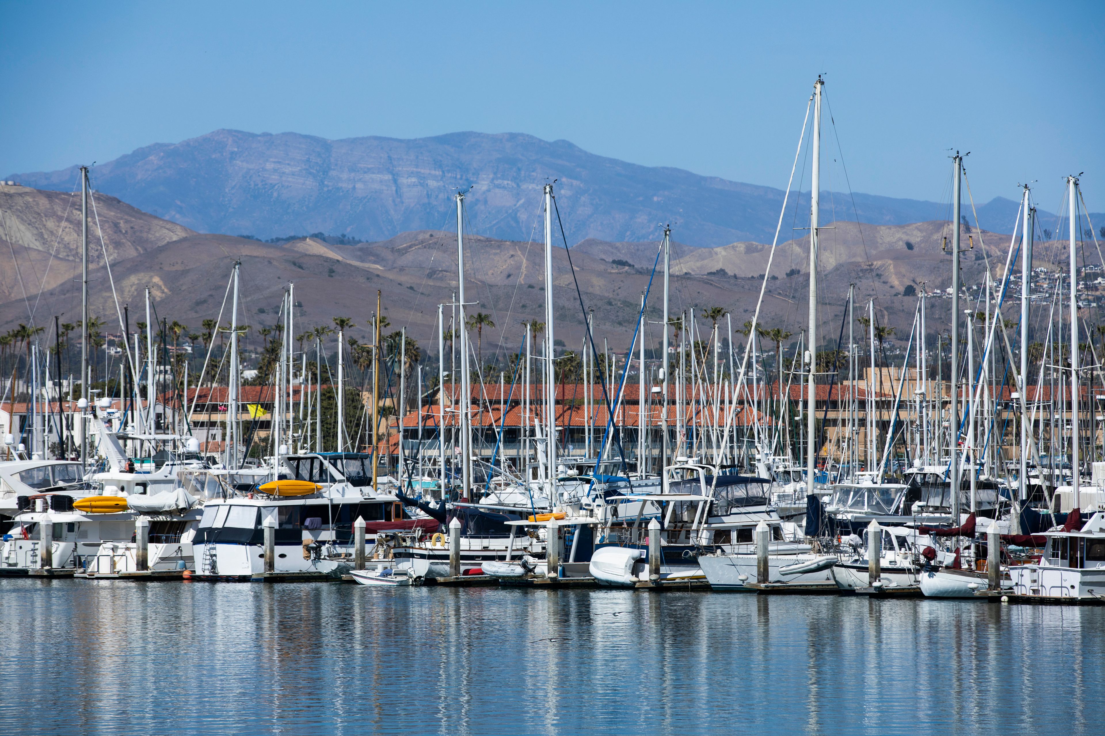 A marina with numerous sailboats docked, set against a backdrop of mountains and a clear sky.