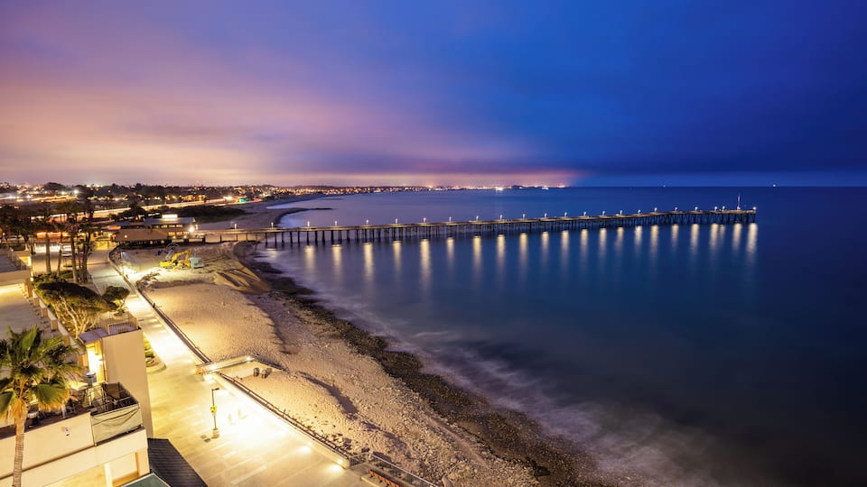 A coastal cityscape at dusk with a long pier extending into the water, a beach, and buildings along the shore.