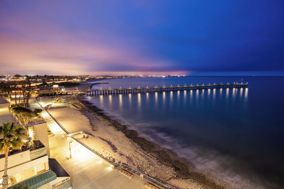 Un paisaje urbano costero al anochecer, con un muelle largo que se adentra en el agua, una playa y edificios a lo largo de la costa.