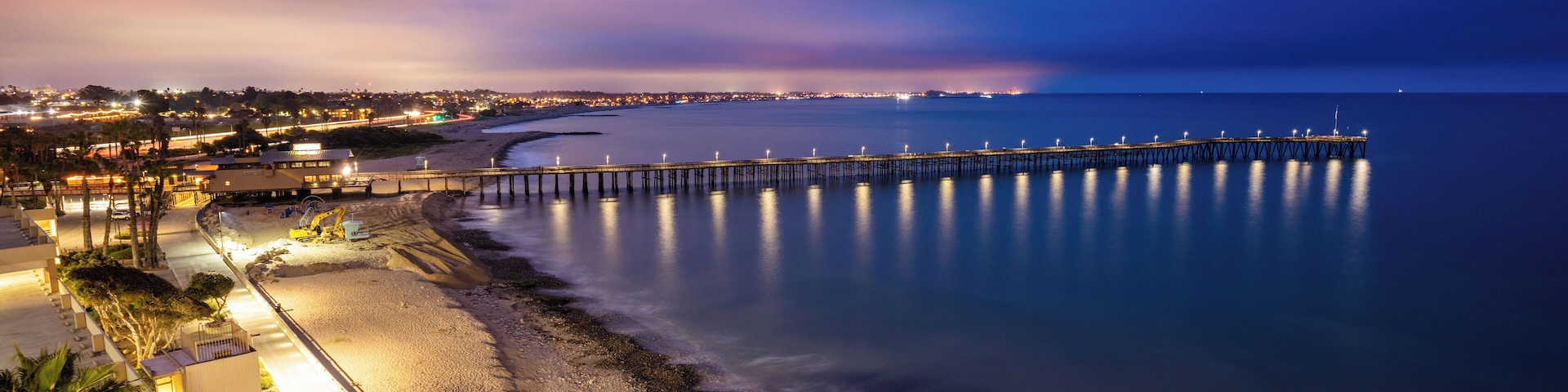 Paisagem costeira ao entardecer, com um longo cais que se estende para a água, uma praia e edifícios ao longo da costa.
