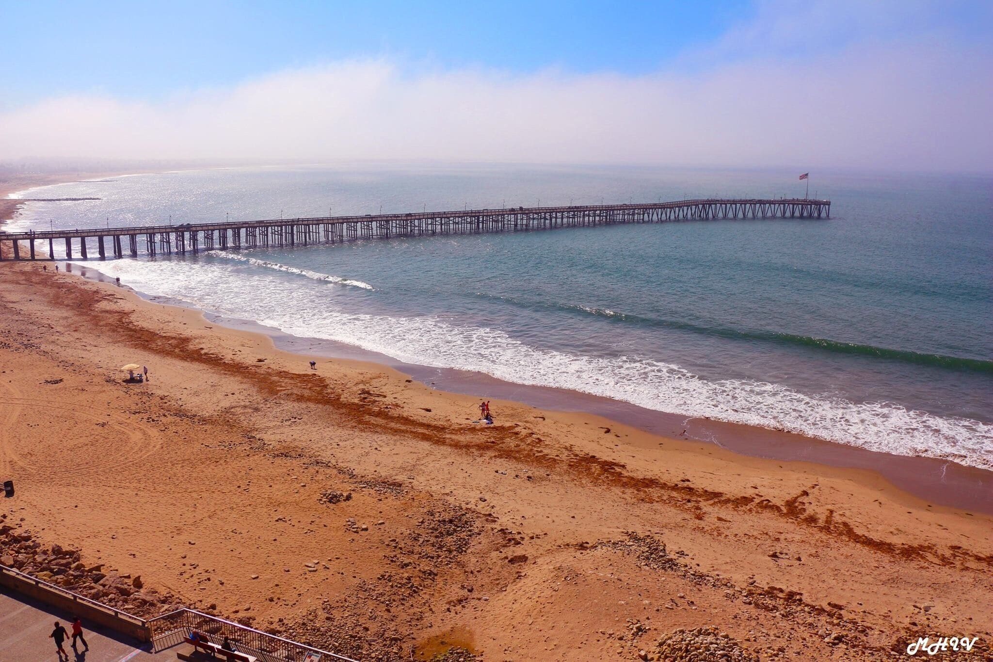 A beach with a long pier extending into the ocean, waves gently lapping at the shore, and a clear sky above.
