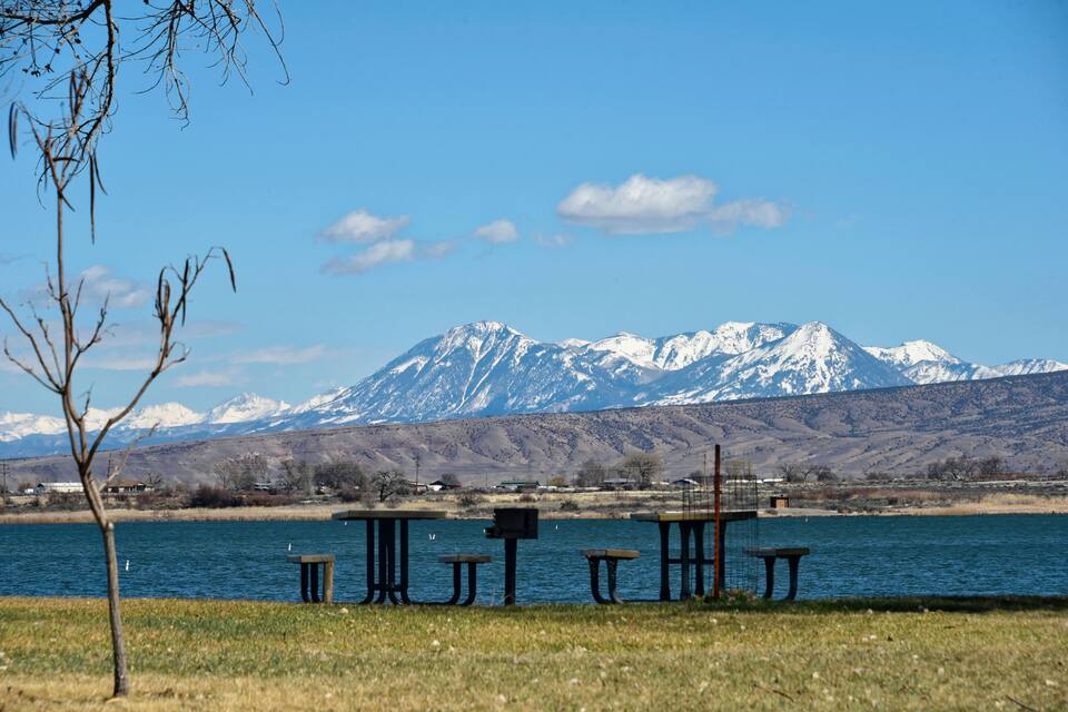A lakeside picnic area with benches and a table, surrounded by a grassy field and a mountain range with snow-capped peaks in the background.
