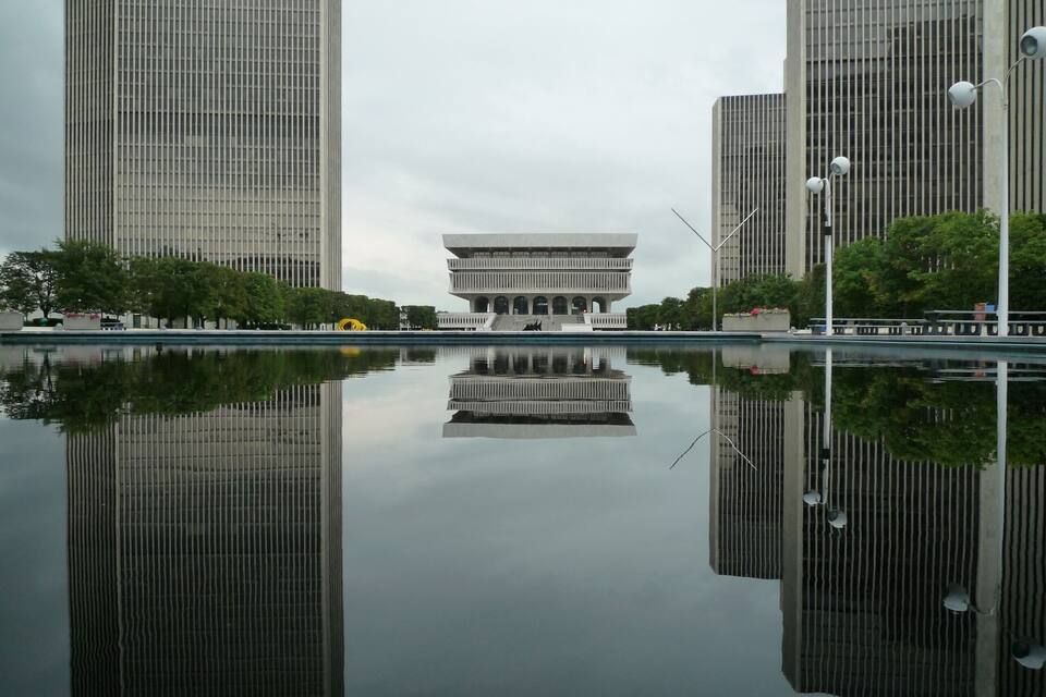 A modern building with a unique design, surrounded by tall skyscrapers and a calm reflective water body.