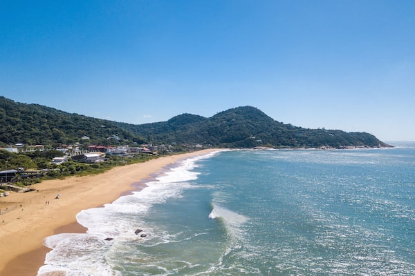A coastal beach with a sandy shore, waves, and a forested hill in the background.