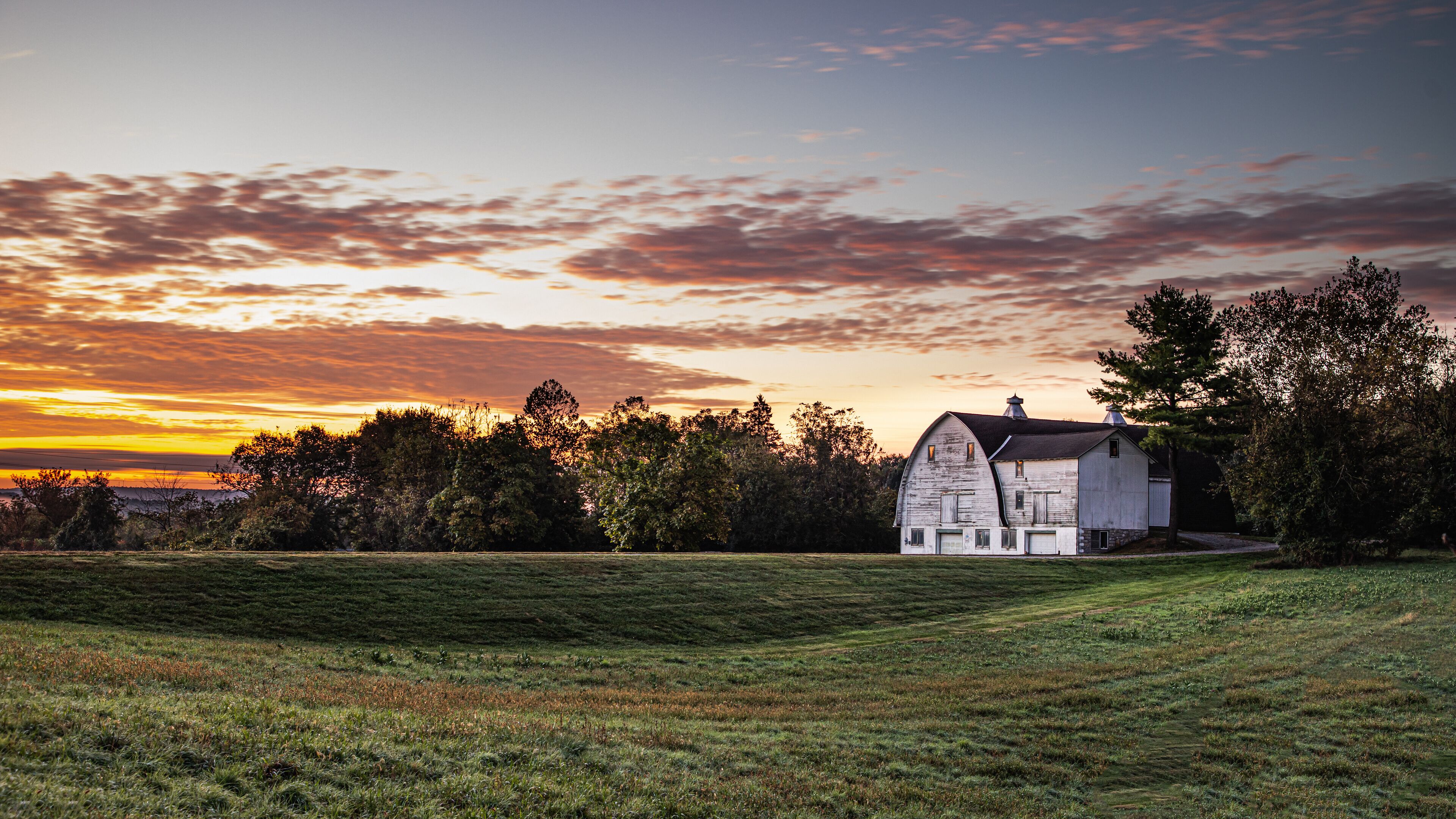 A white barn with a sloped roof in a grassy field, surrounded by trees and a sunset sky.