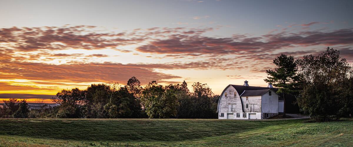 A white barn with a sloped roof in a grassy field, surrounded by trees and a sunset sky.