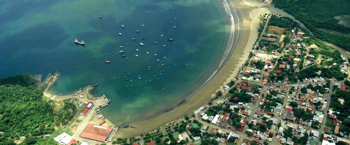 An aerial view of a coastal town with a beach, boats, and dense greenery.