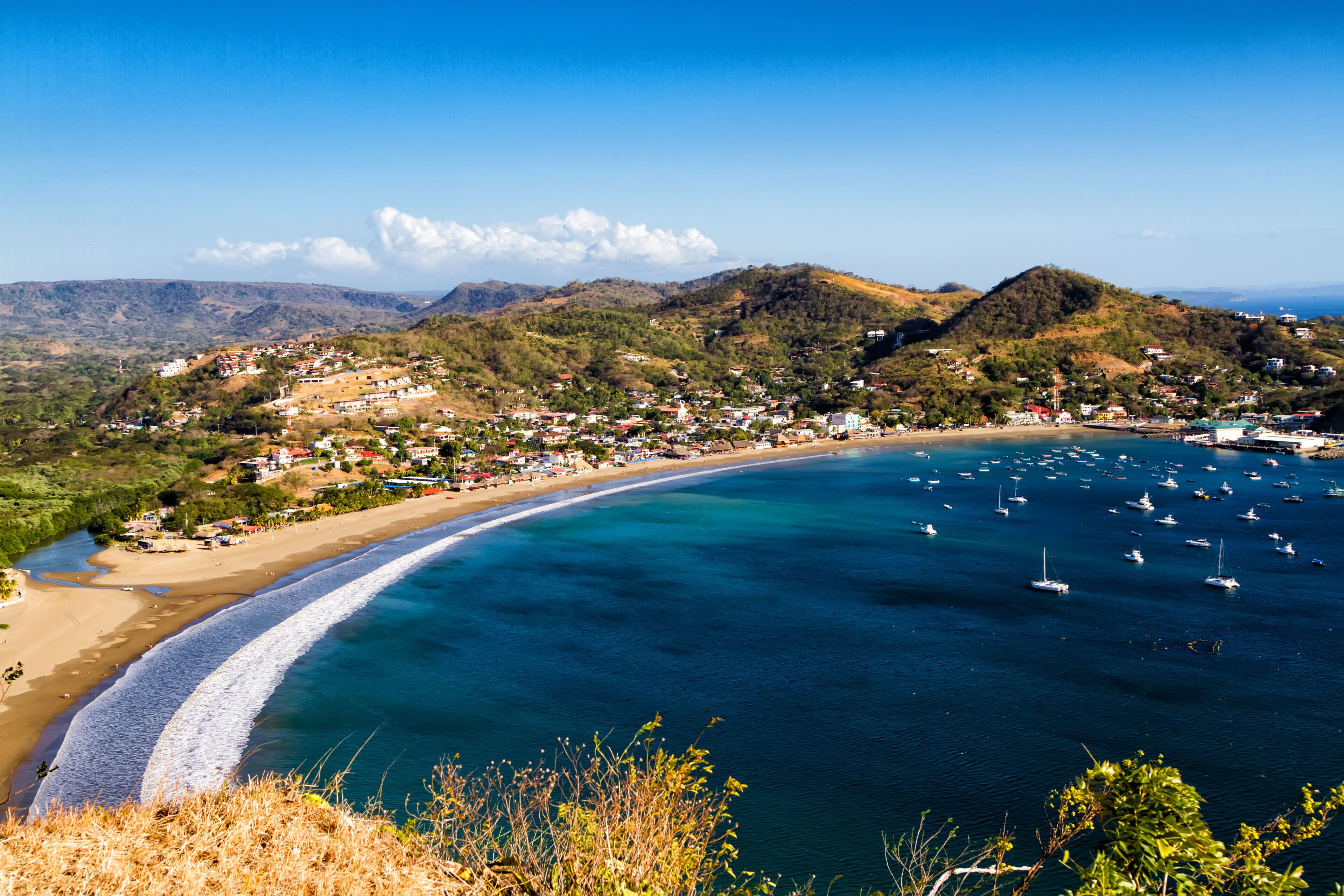 Uma cidade costeira com praia, águas azul-claras, barcos e colinas ao fundo.