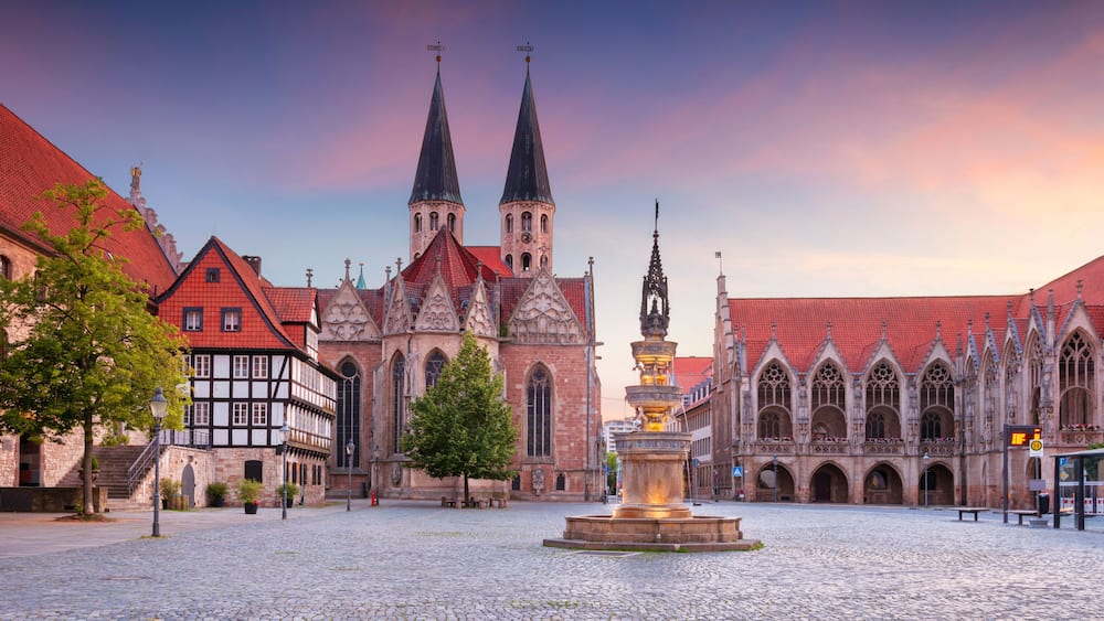A historic square with a prominent church featuring twin spires, a fountain, and traditional half-timbered buildings.
