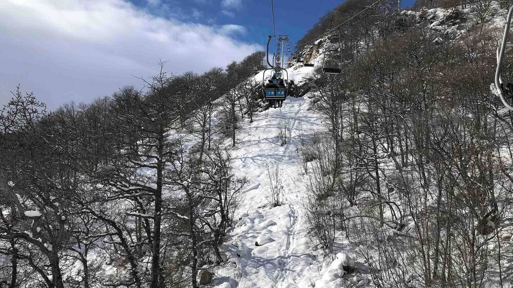 A ski lift ascending a snow-covered mountain with trees and rocky terrain.