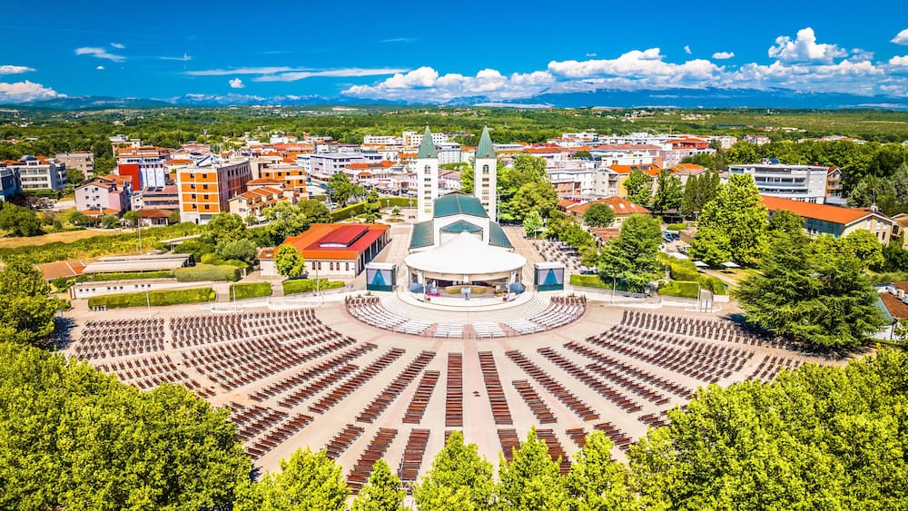Drone image of Medjugorje showing central church, large open square, surrounding buildings and visitors gathered at this Christian pilgrimage site under clear skies in summer