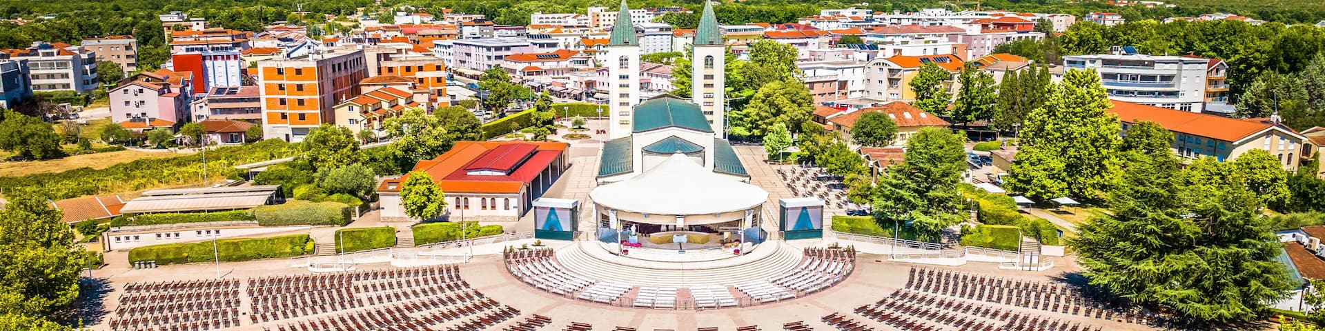 Drone image of Medjugorje showing central church, large open square, surrounding buildings and visitors gathered at this Christian pilgrimage site under clear skies in summer