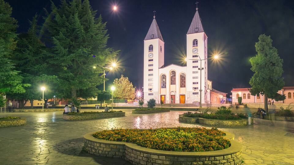 A church with two towers at night, surrounded by a courtyard with a fountain and trees.