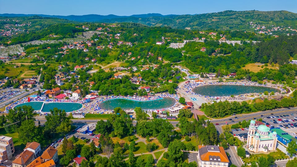 An aerial view of a coastal town with two large swimming pools, a beach, and a church with domes.