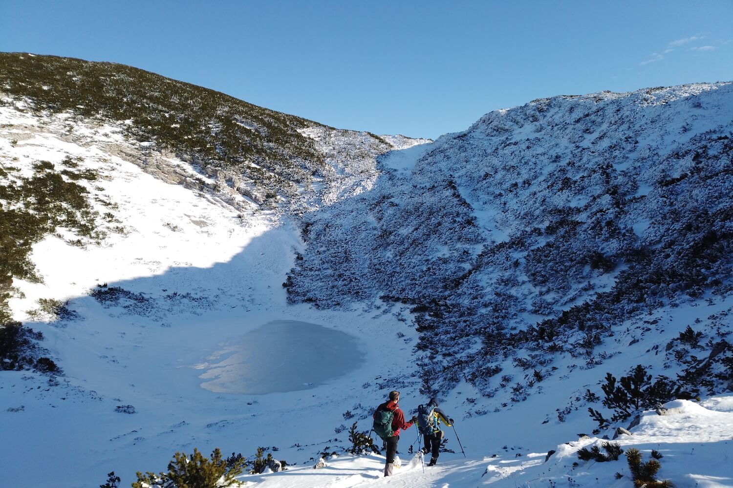 A snowy mountain landscape with a small lake and two hikers.