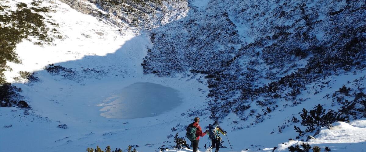 A snowy mountain landscape with a small lake and two hikers.