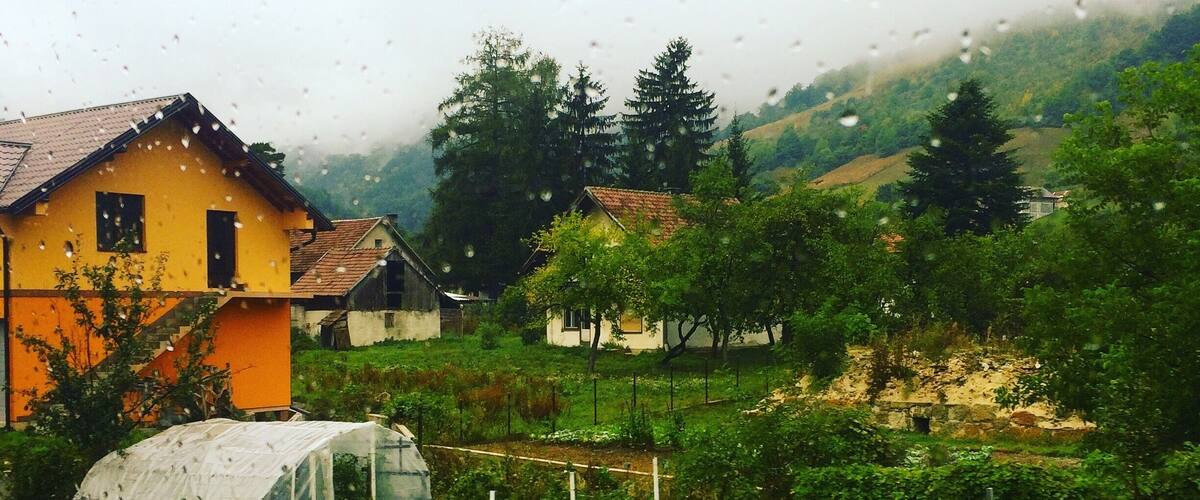 A rural landscape with a yellow house, a greenhouse, and a vegetable garden.