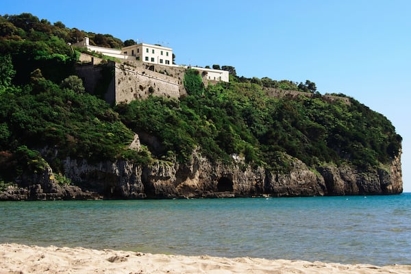 Falésias costeiras com um edifício no topo, uma praia de areia e água azul cristalina.