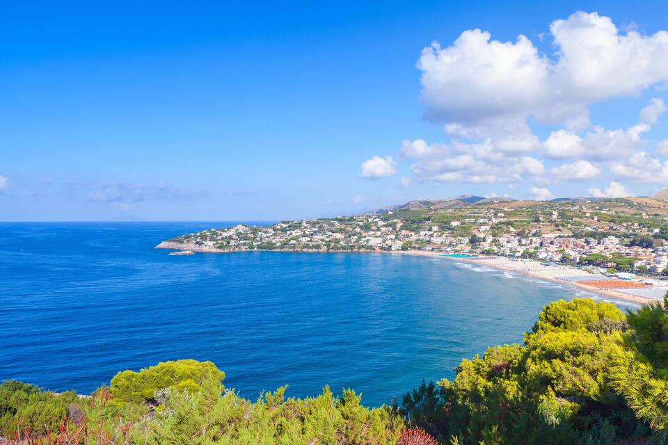 A coastal town with a beach, clear blue sea, and green hills.