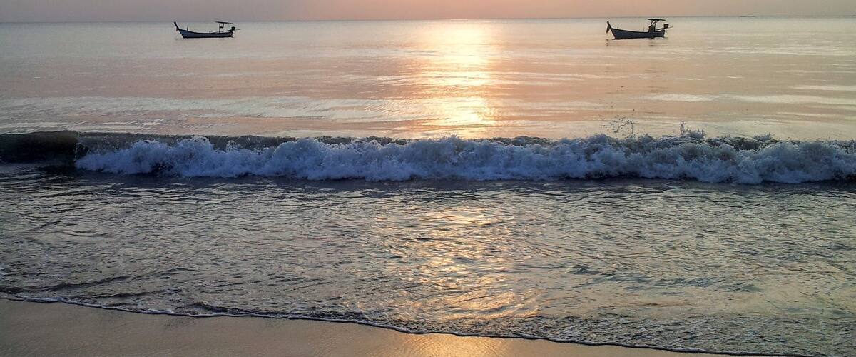 A beach with waves at sunset, two boats in the distance, and a clear sky.