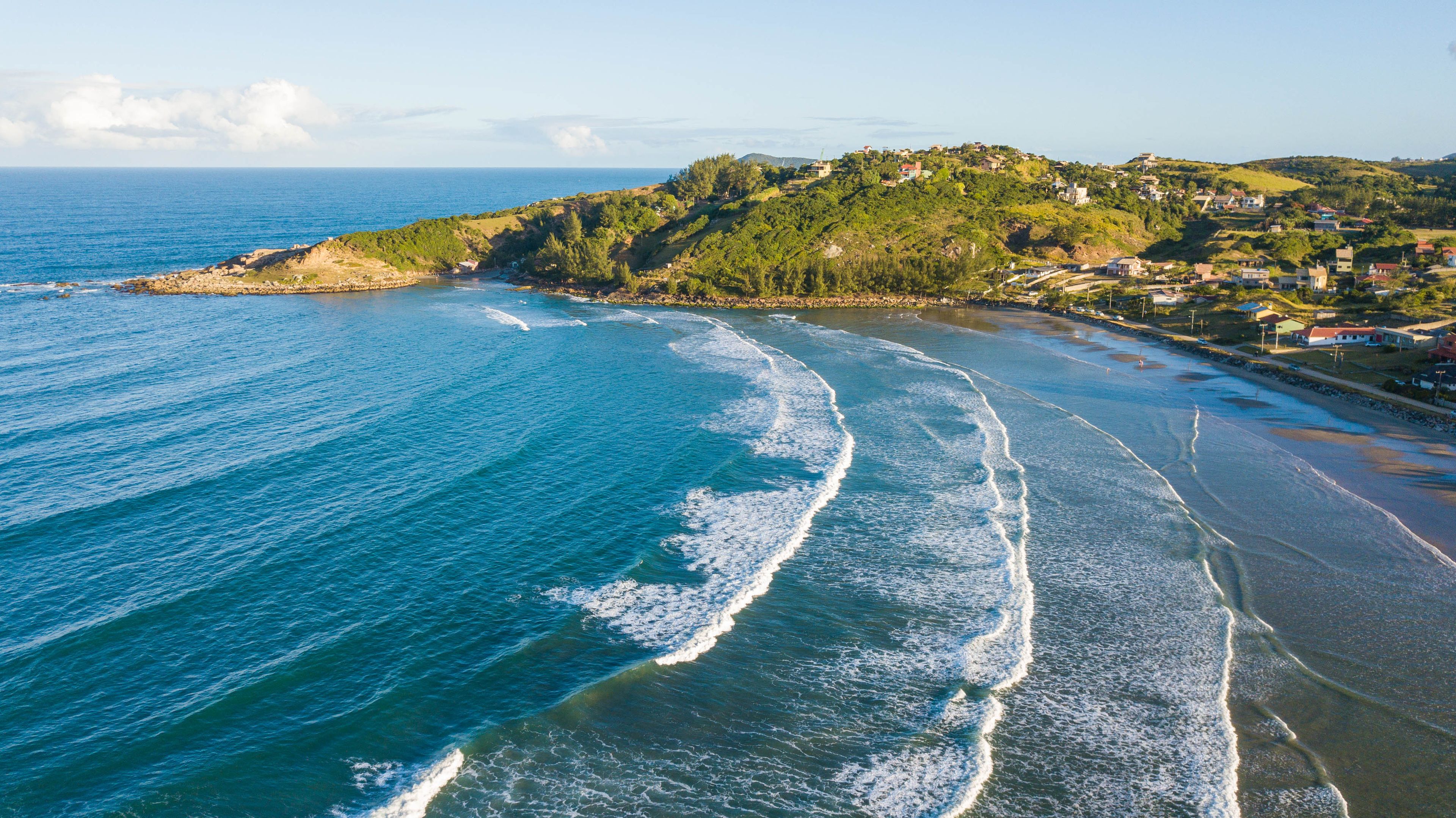 A coastal landscape with a beach, waves, and a hillside with houses.