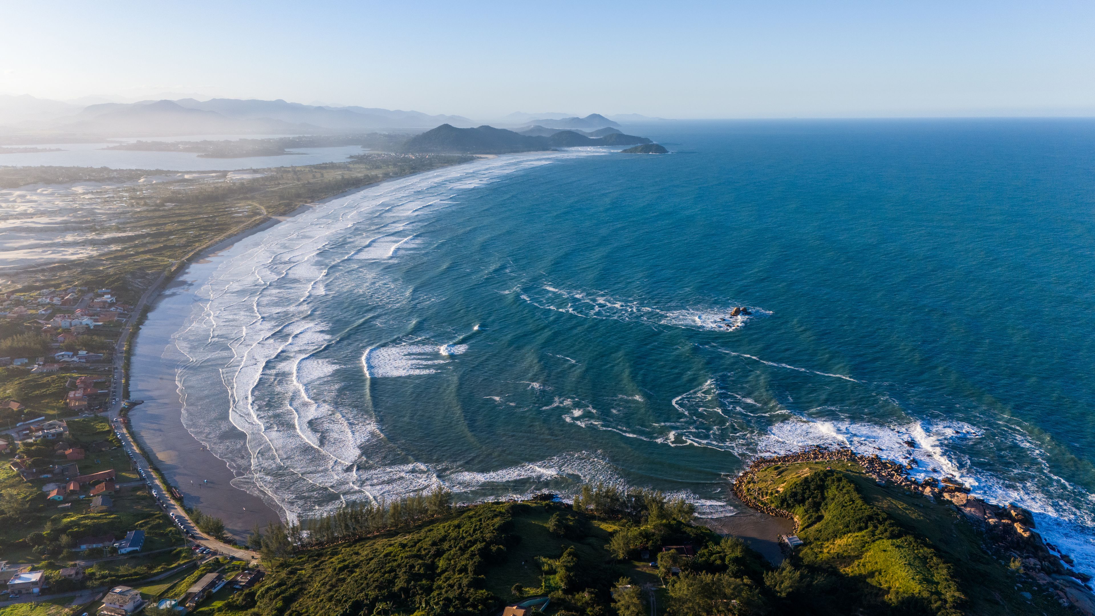 An aerial view of a coastal area with a beach, ocean waves, and a distant mountain range.