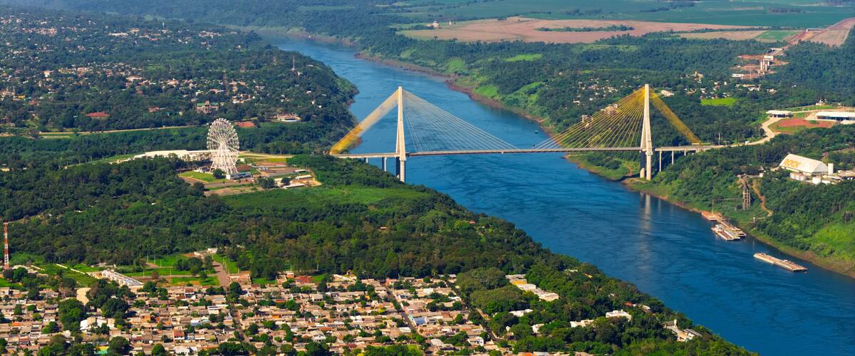 A large suspension bridge over a river, surrounded by lush greenery and a small town.