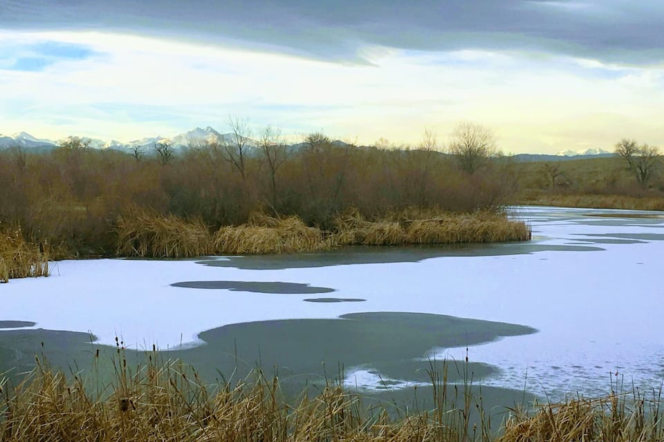 A frozen river with patches of ice, surrounded by dry grass and trees under a cloudy sky.