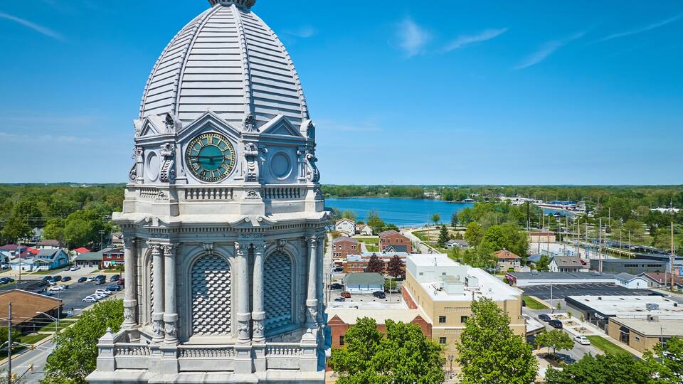 A clock tower with a dome and Roman numerals, overlooking a town with a lake in the background.