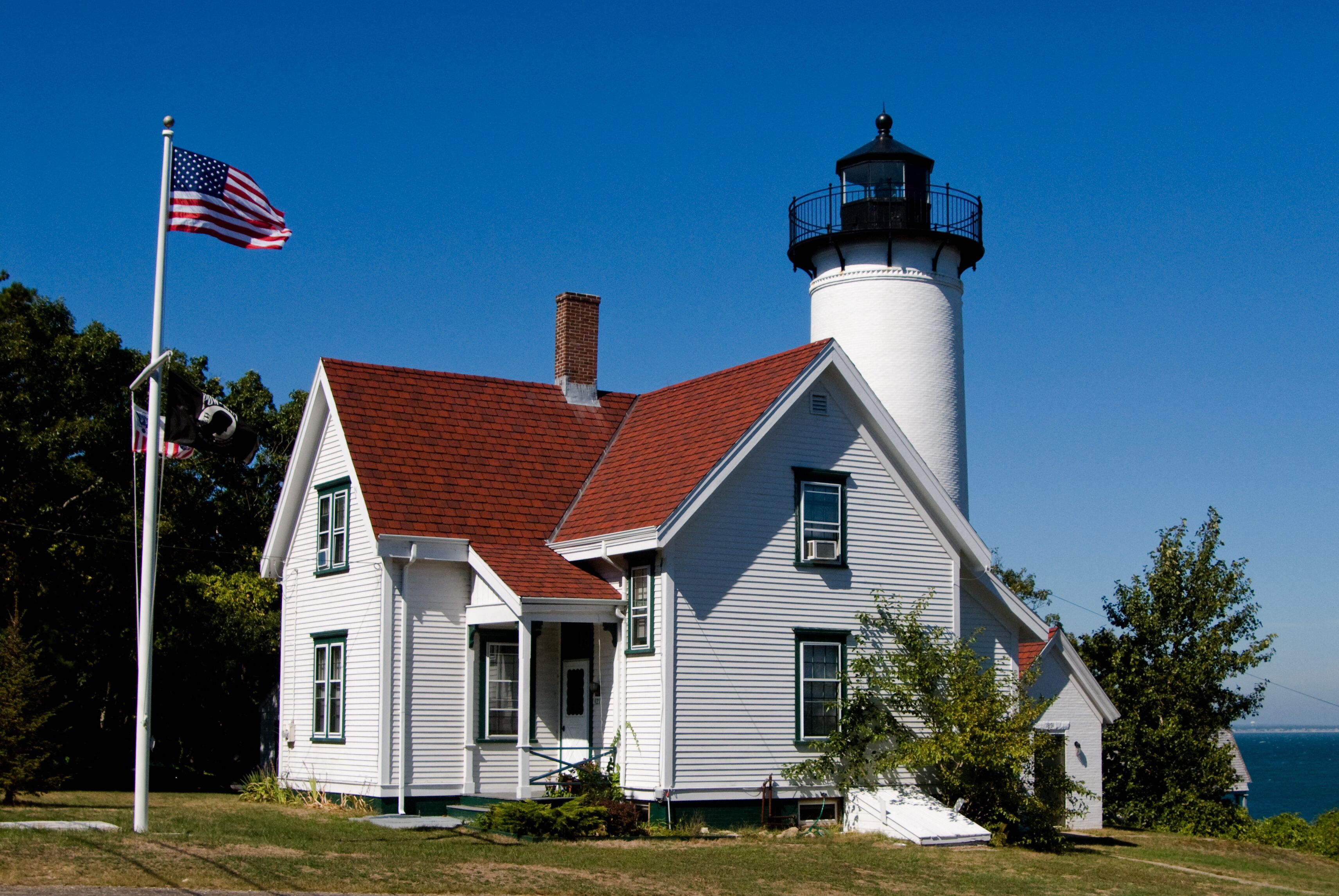 A white lighthouse with a red roof, an American flag, and a view of the ocean.